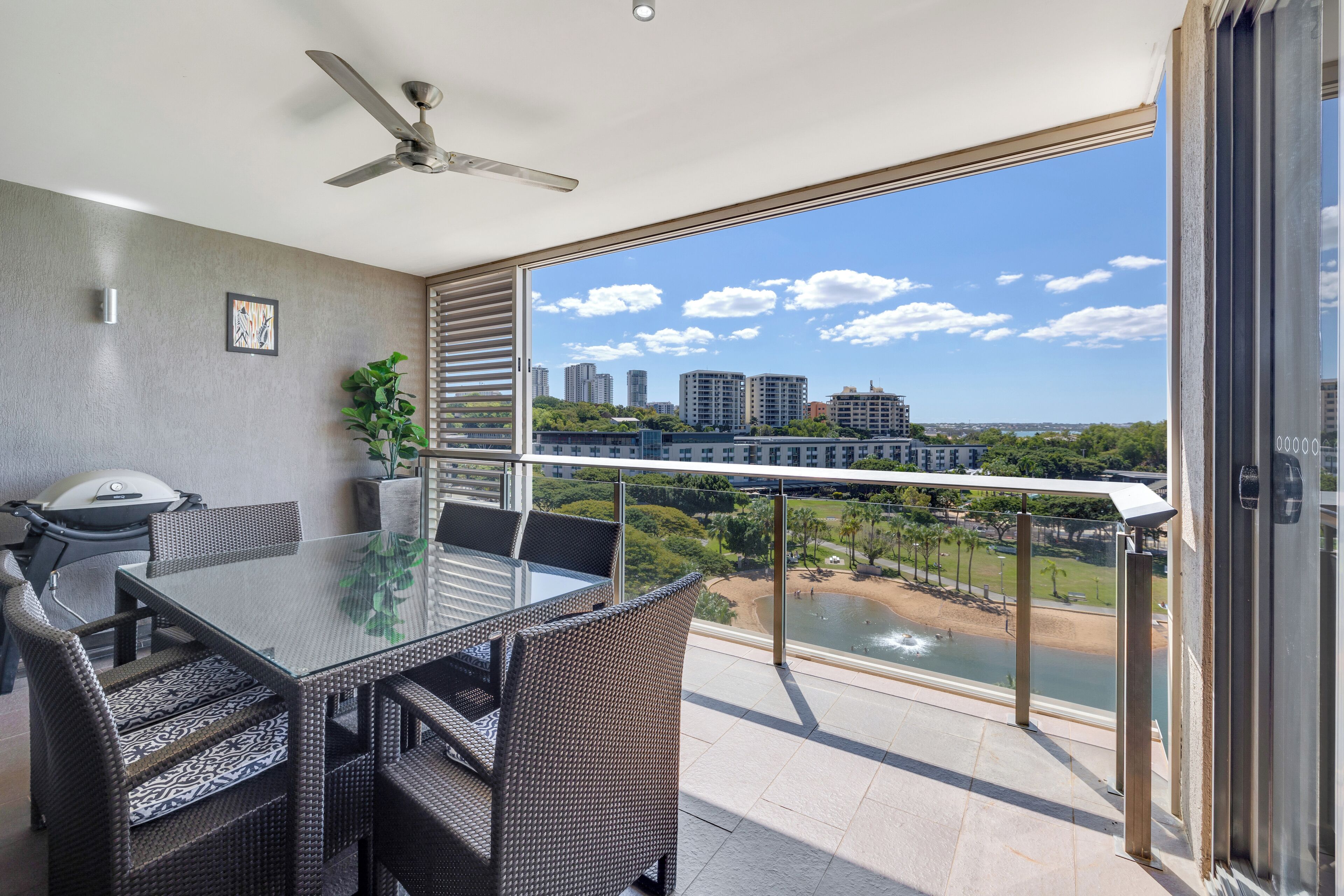 A balcony with a glass table and chairs, overlooking a beach and cityscape.