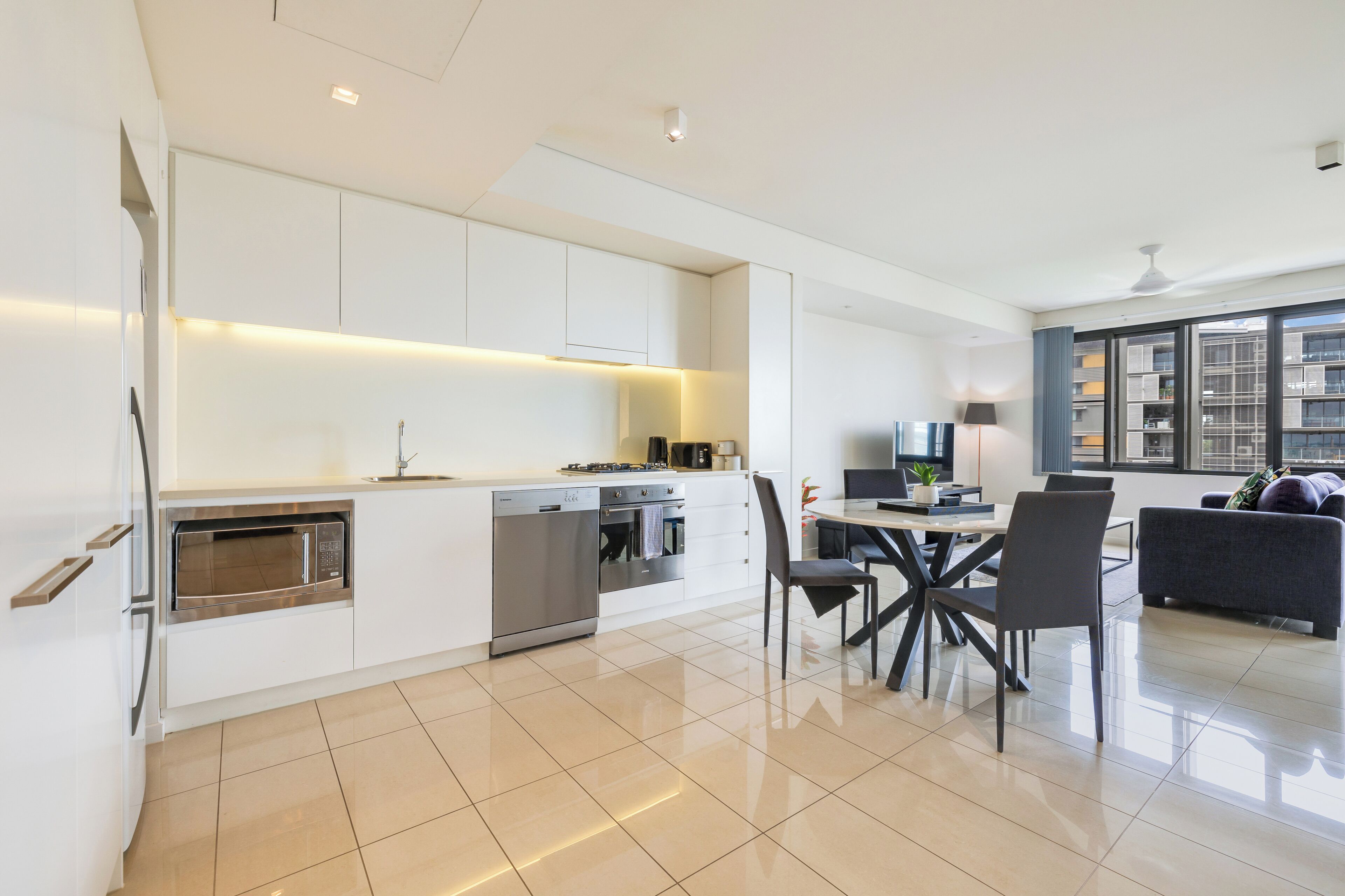Une cuisine moderne avec des meubles blancs, un coin repas comprenant une table et des chaises, et un salon avec un canapé et une grande fenêtre.