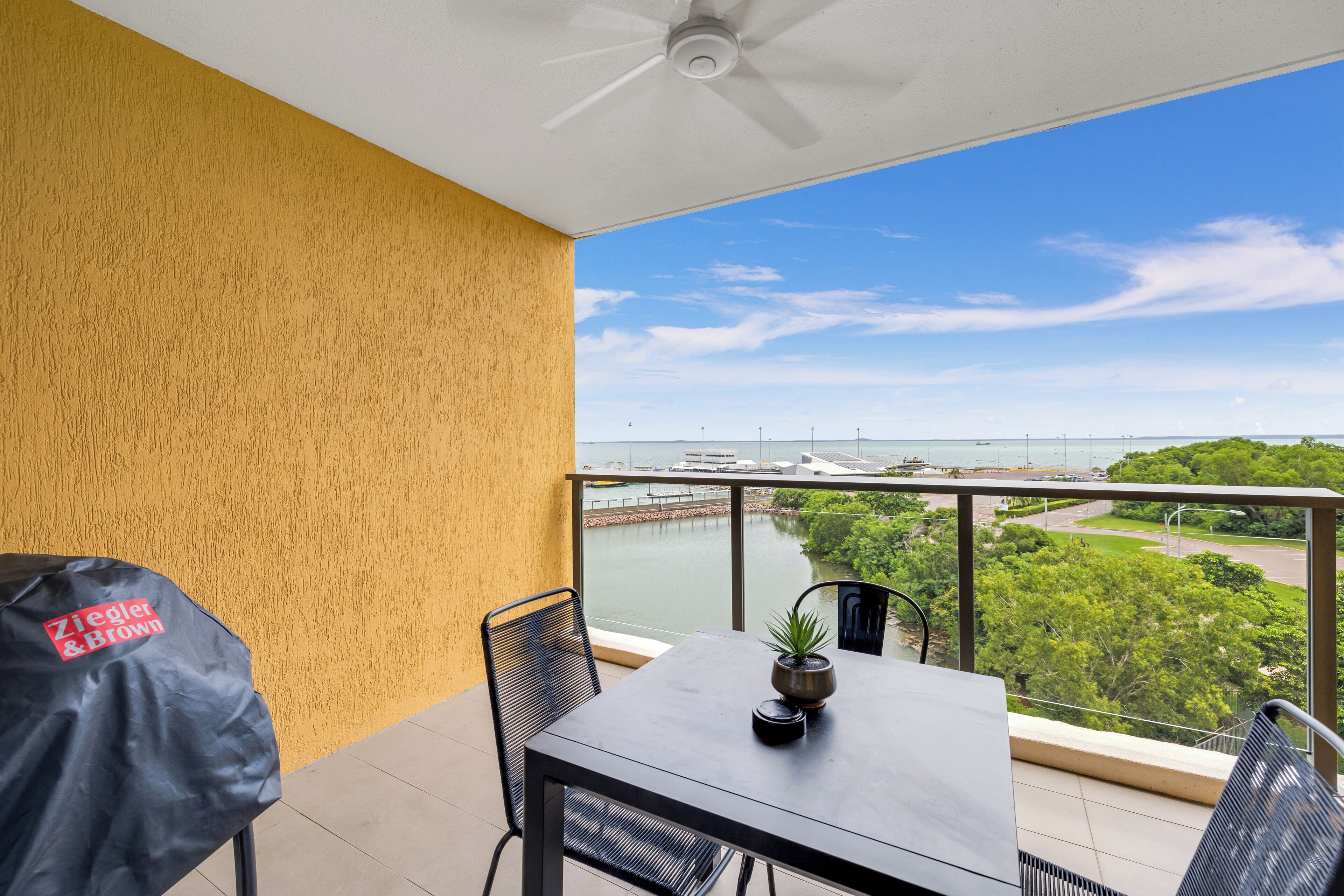 A balcony with a view of a river and bridge, a table with chairs, and a ceiling fan.