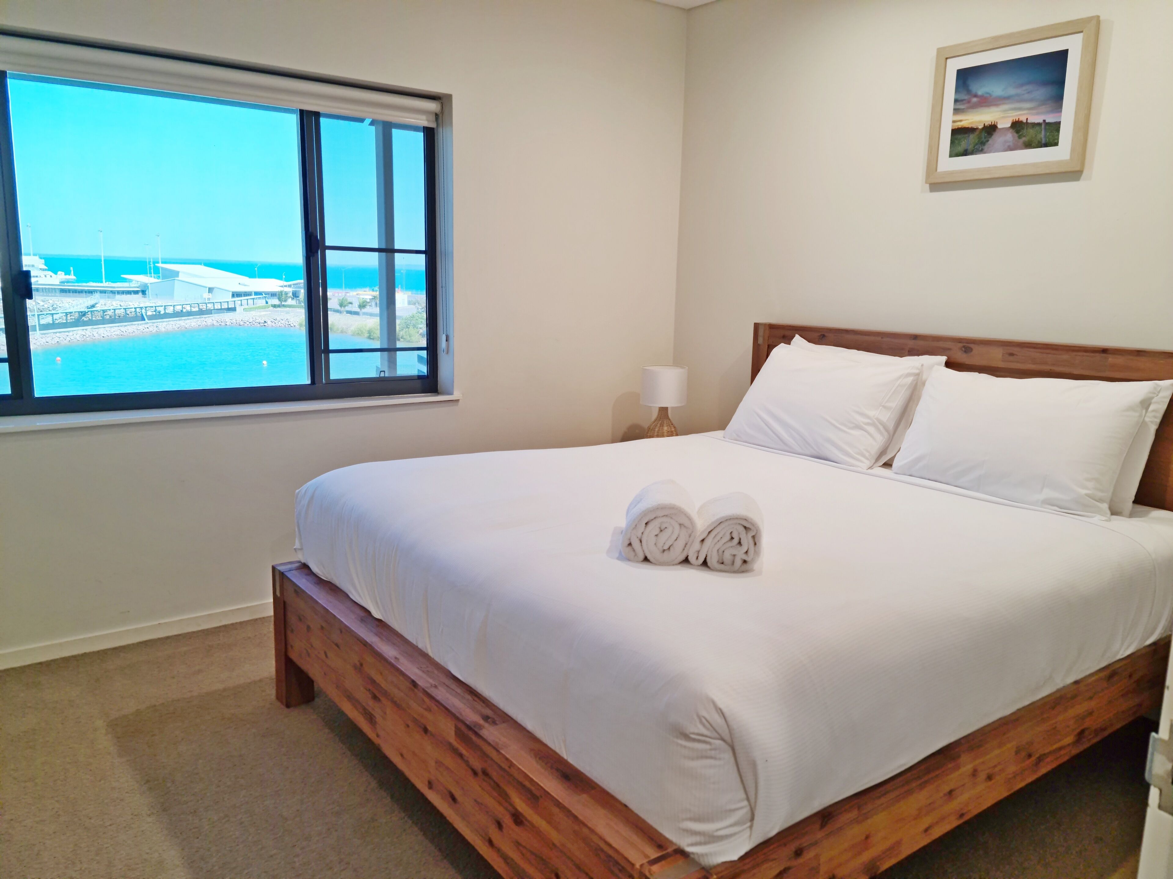 A neatly made bed with white linens and two rolled towels, a wooden headboard, a nightstand with a lamp, a framed picture on the wall, and a window with a view of a marina and boats.