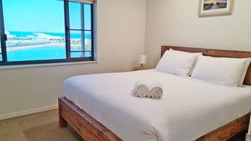 A neatly made bed with white linens and two rolled towels, a wooden headboard, a nightstand with a lamp, a framed picture on the wall, and a window with a view of a marina and boats.
