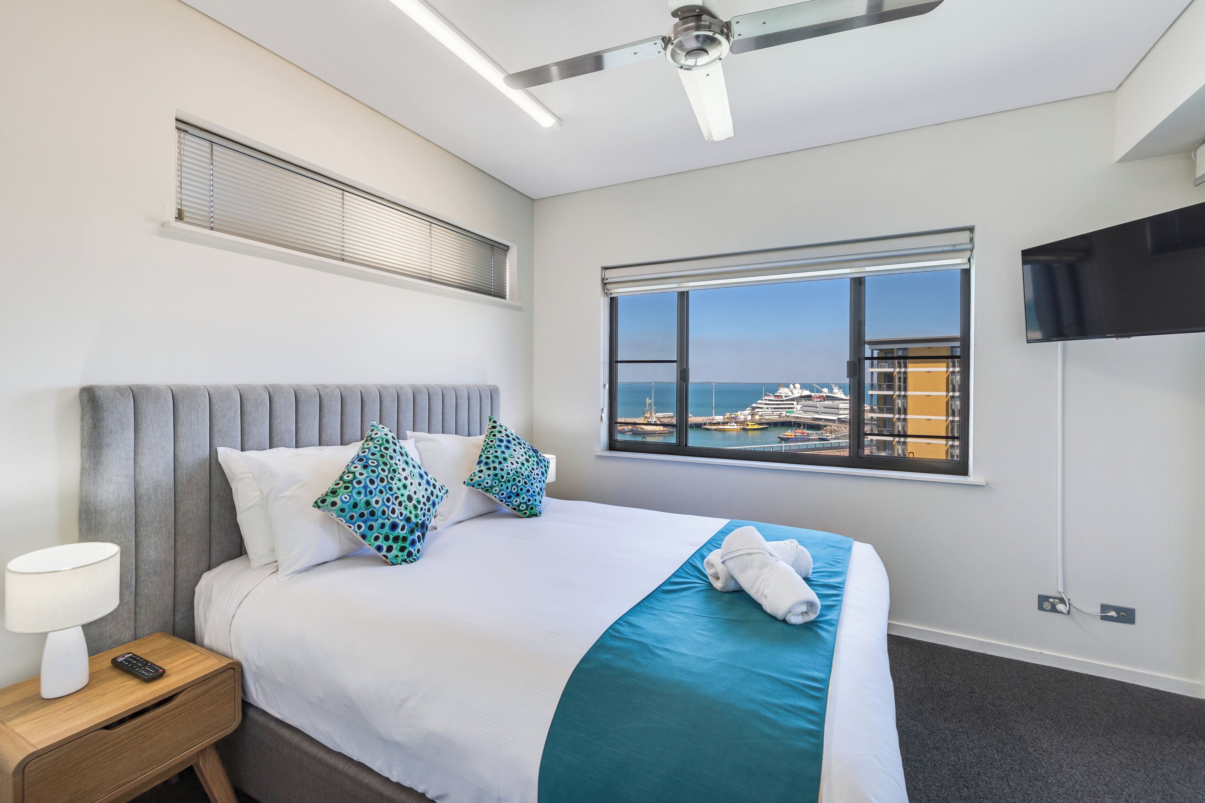 Une chambre d’hôtel comprenant un lit, une table de chevet, une télécommande, un ventilateur de plafond, une fenêtre donnant sur la mer et un téléviseur fixé au mur.