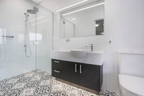 A modern bathroom with a glass shower enclosure, a white sink, dark cabinets, and patterned floor tiles. - The Apartment, Metung (Metung)