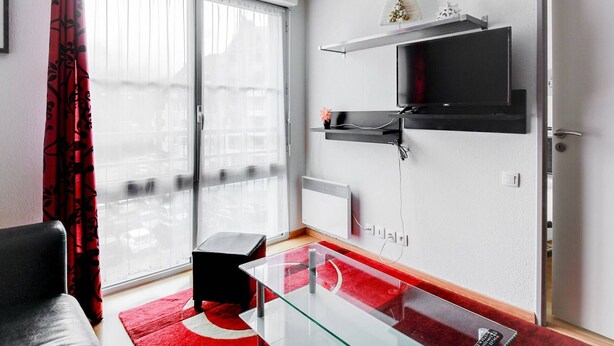 Un salón moderno con una mesa de centro de vidrio, un sofá negro, una alfombra roja y blanca, un televisor montado en la pared y un ventanal corredero que da a un balcón.