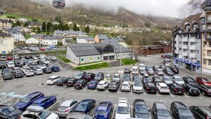 Un estacionamiento con varios autos, un edificio con gran fachada de vidrio y un paisaje montañoso con un teleférico a lo lejos.