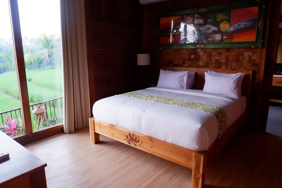 A wooden bed with white bedding, a nightstand, a lamp, and a view of a green landscape through large windows.