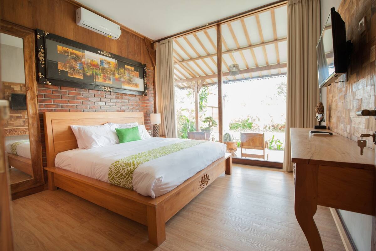 A wooden bed with white and green bedding, a wooden nightstand, a TV mounted on the wall, and a large window with a view of greenery.