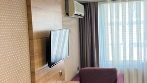 A hotel room with a wooden wall panel, a flat-screen TV mounted above a wooden table, a purple sofa, and a window with sheer curtains. - Prens Hotel (Istanbul)