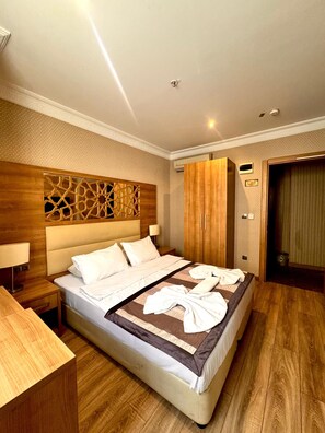 A hotel room with a large bed, wooden headboard, and a decorative wall panel. There is a nightstand with a lamp, a wooden desk, and a door with a sign indicating no smoking. - Prens Hotel (Istanbul)