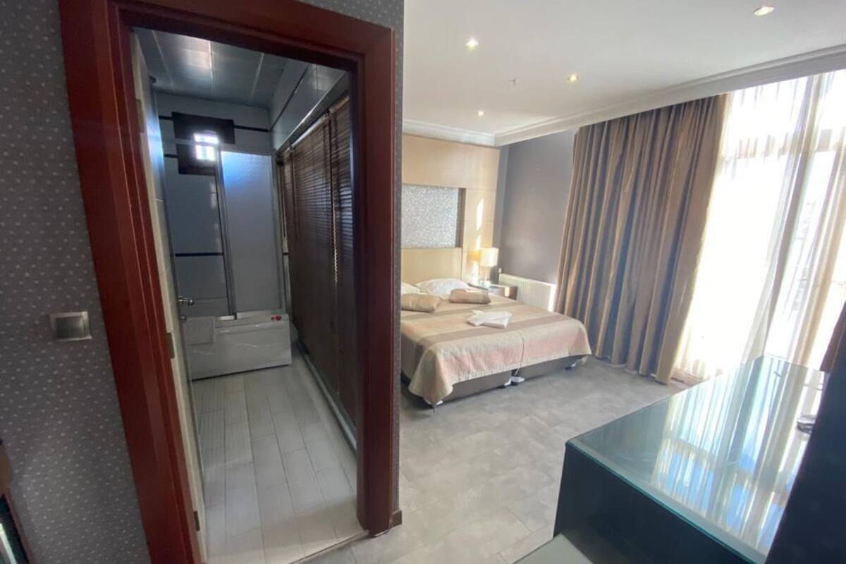 A hotel room with a bed, a bedside table, a lamp, a window with curtains, and a view of the hallway.
