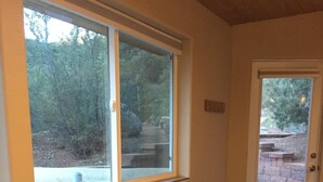 A beige sofa with patterned cushions, a wooden chair, and a window with a view of trees and a brick wall. - Casita Canoncito--private suite with kitchenette (ALbuquerque)