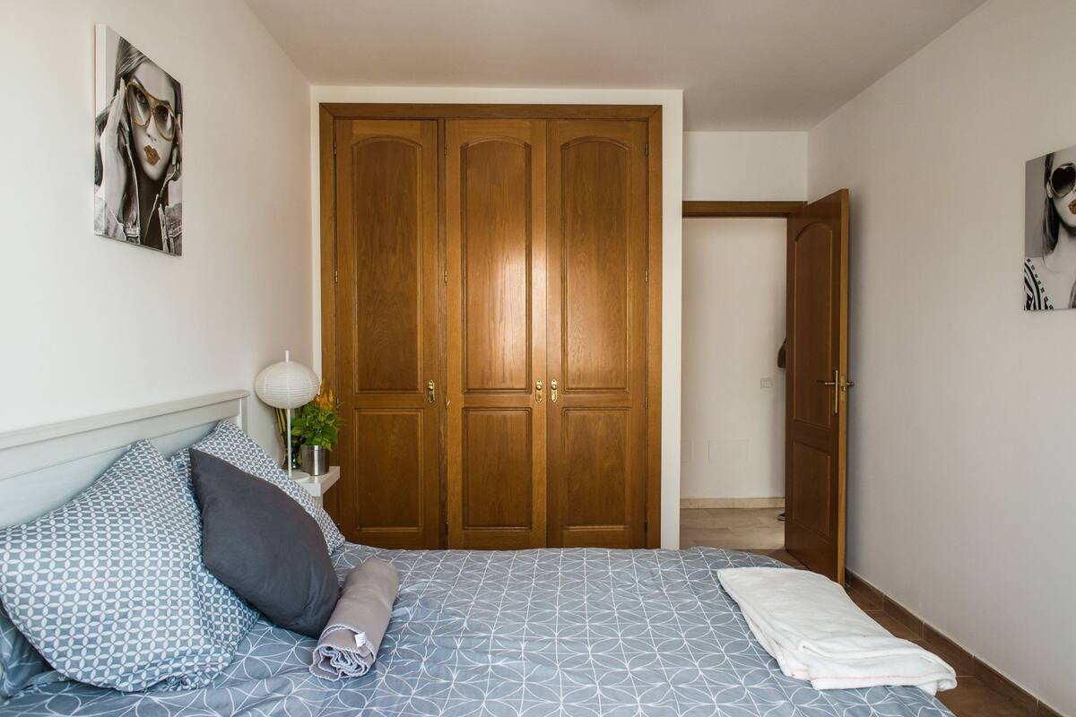 Une chambre avec une grande armoire en bois, un lit avec une literie à motifs et une table de chevet avec une lampe et une plante.