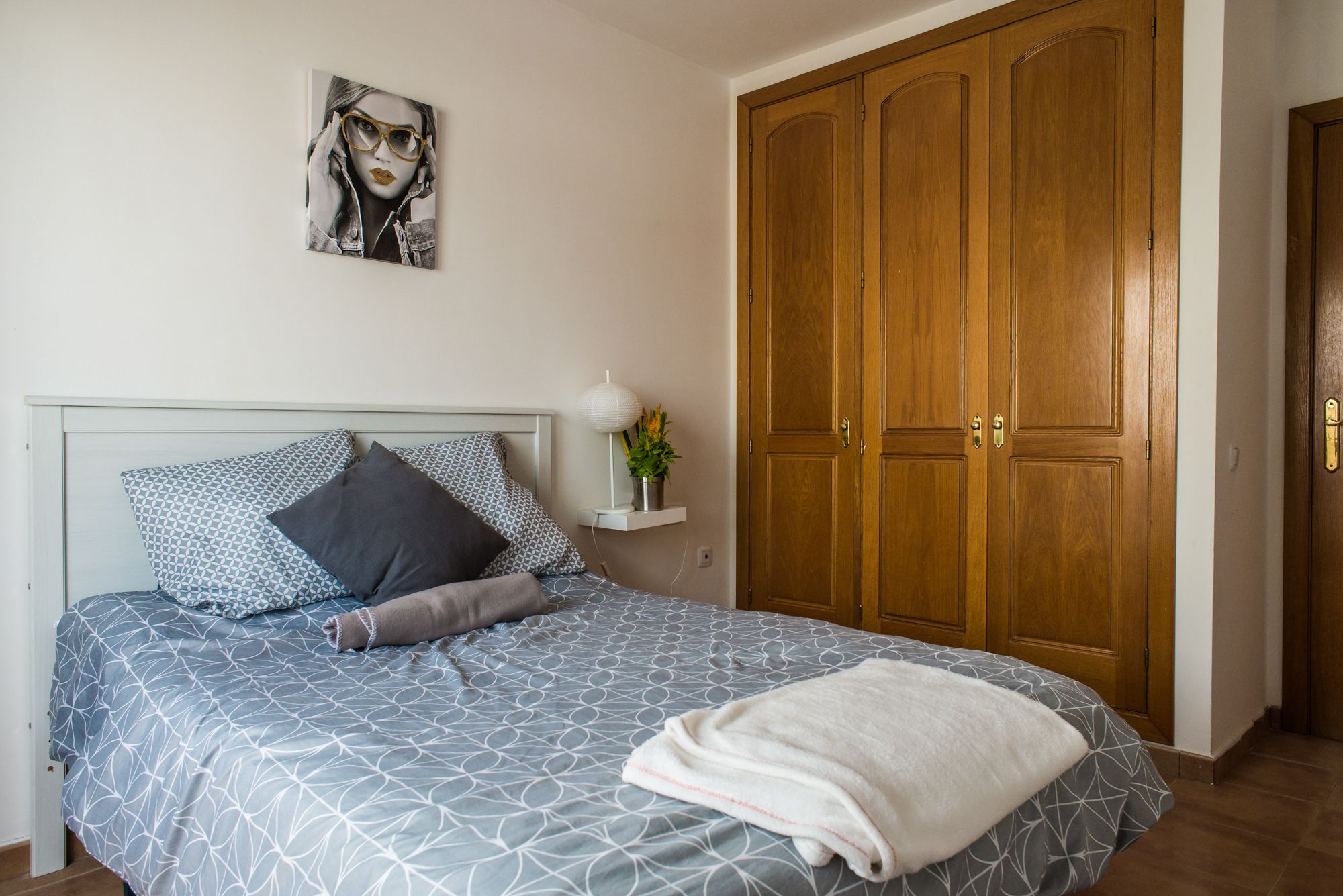 Un dormitorio con cama, almohadas, una mesita de noche con lámpara y planta, y un armario de madera.
