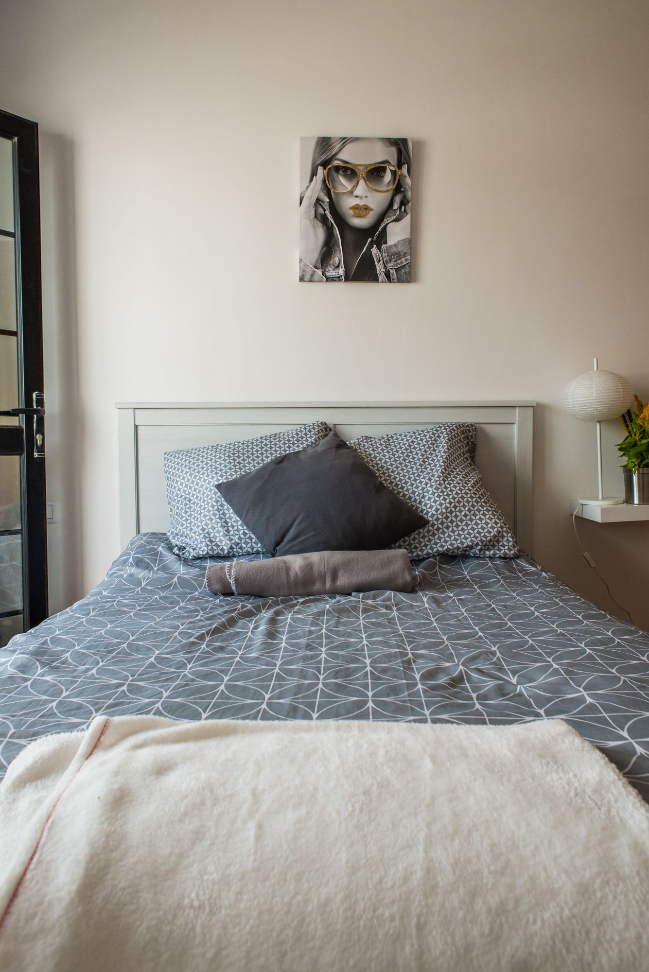 Een net opgemaakt bed met beddengoed met een patroon, een grijze en witte kussenset, en een kunstwerk in een lijst erboven.