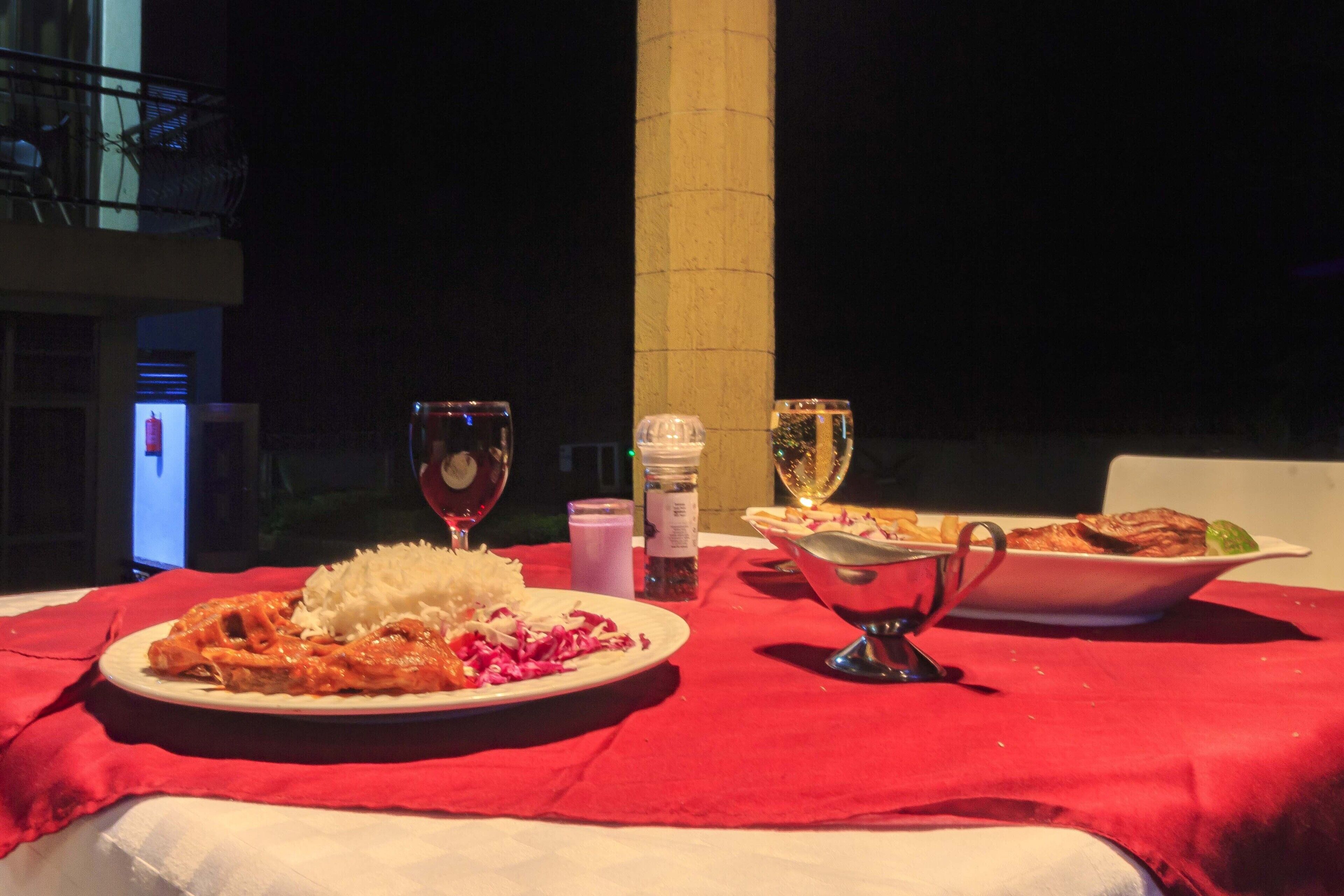 A table set with a red tablecloth, featuring a plate of food, two glasses of wine, and condiments.