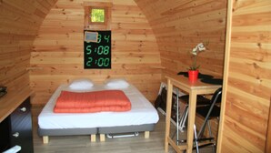 Ein Zimmer in einer Holzhütte mit einem Bett, einem kleinen Tisch und einer digitalen Uhr, die die Zeit anzeigt.