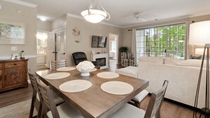 A dining area with a wooden table, chairs, and a chandelier. There is a sofa and a television in the background. - River Crossing #1224 2 Bedroom Condo by RedAwning (North Myrtle Beach)