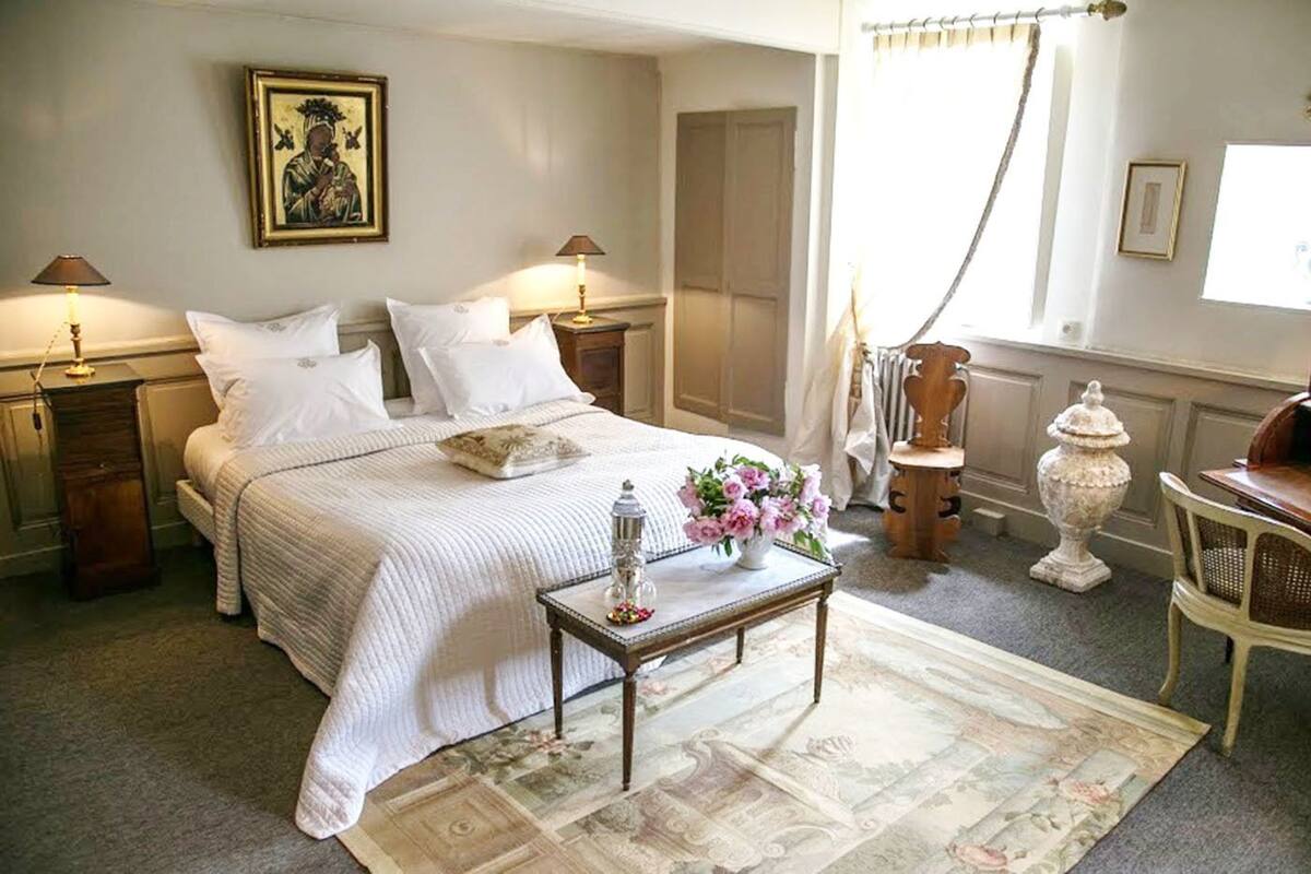 A neatly arranged bedroom with a large bed, bedside tables, a small table with flowers, and a decorative urn.