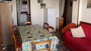A compact living space with a dining table, chairs, a red sofa, a small kitchen area with a refrigerator, and a clock on the wall. - Villa Dilva (San Michele al Tagliamento)