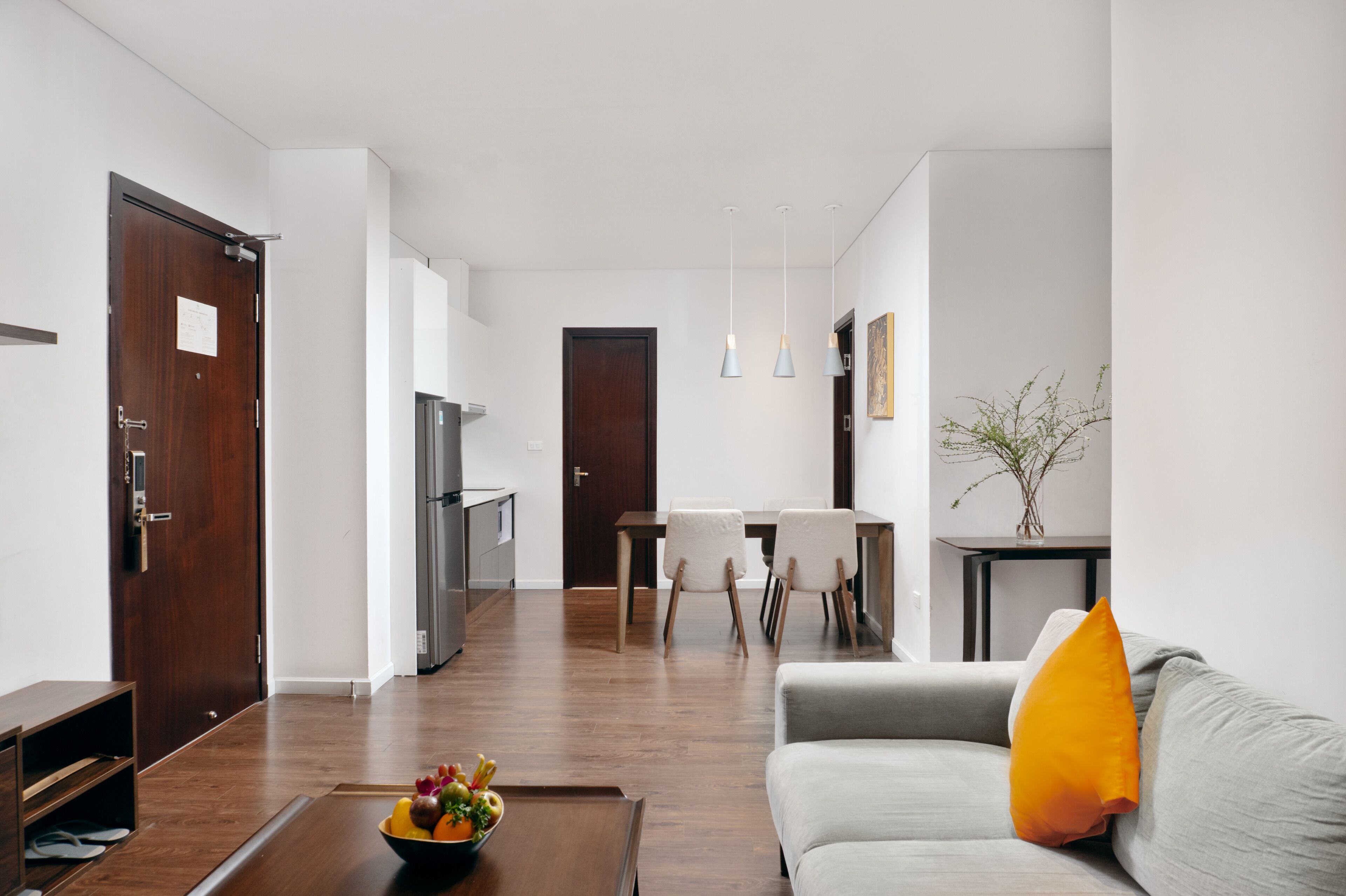 Una sala de estar moderna con un sofá gris, una mesa de centro de madera con fruta, un comedor con sillas y una cocina al fondo.