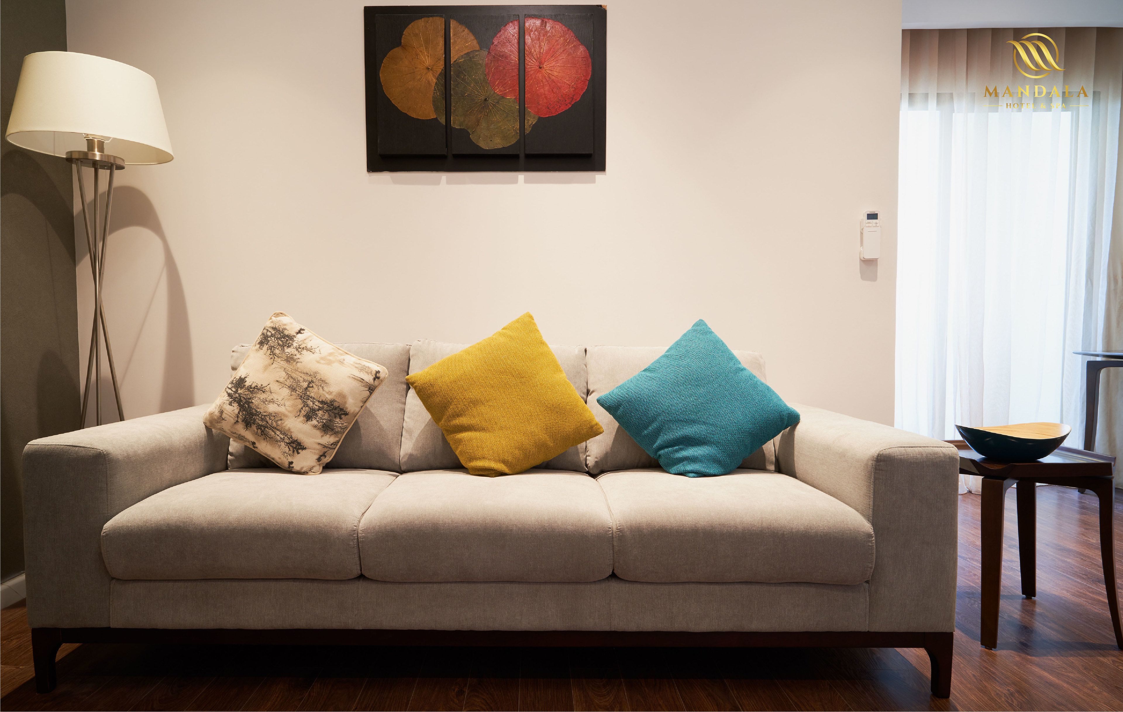 A beige sofa with three pillows in different colors, a floor lamp, a painting on the wall, and a window with curtains.