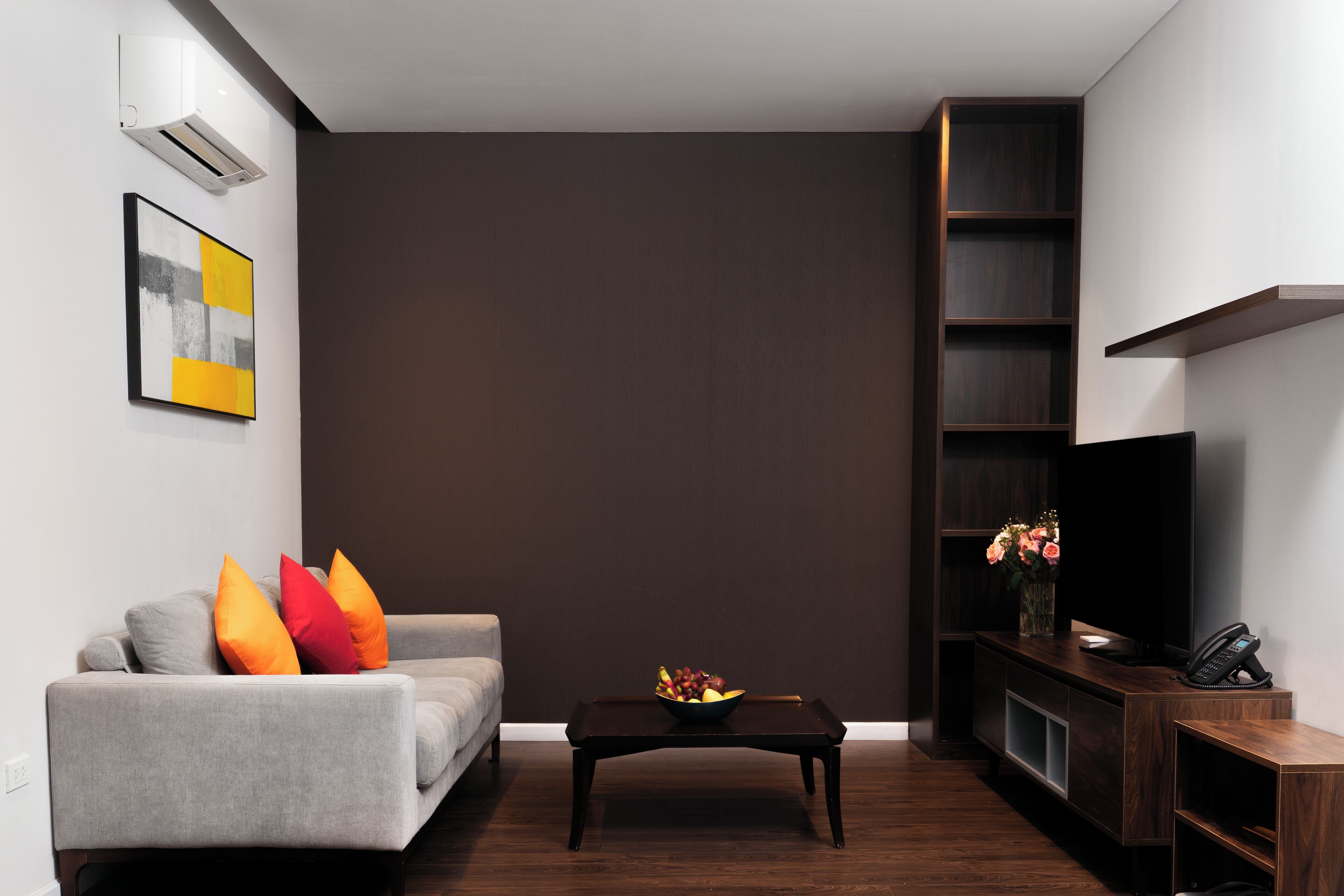 Una sala de estar moderna con un sofá gris, cojines coloridos, una mesa de centro con fruta, un televisor de pantalla plana y una estantería de madera oscura.