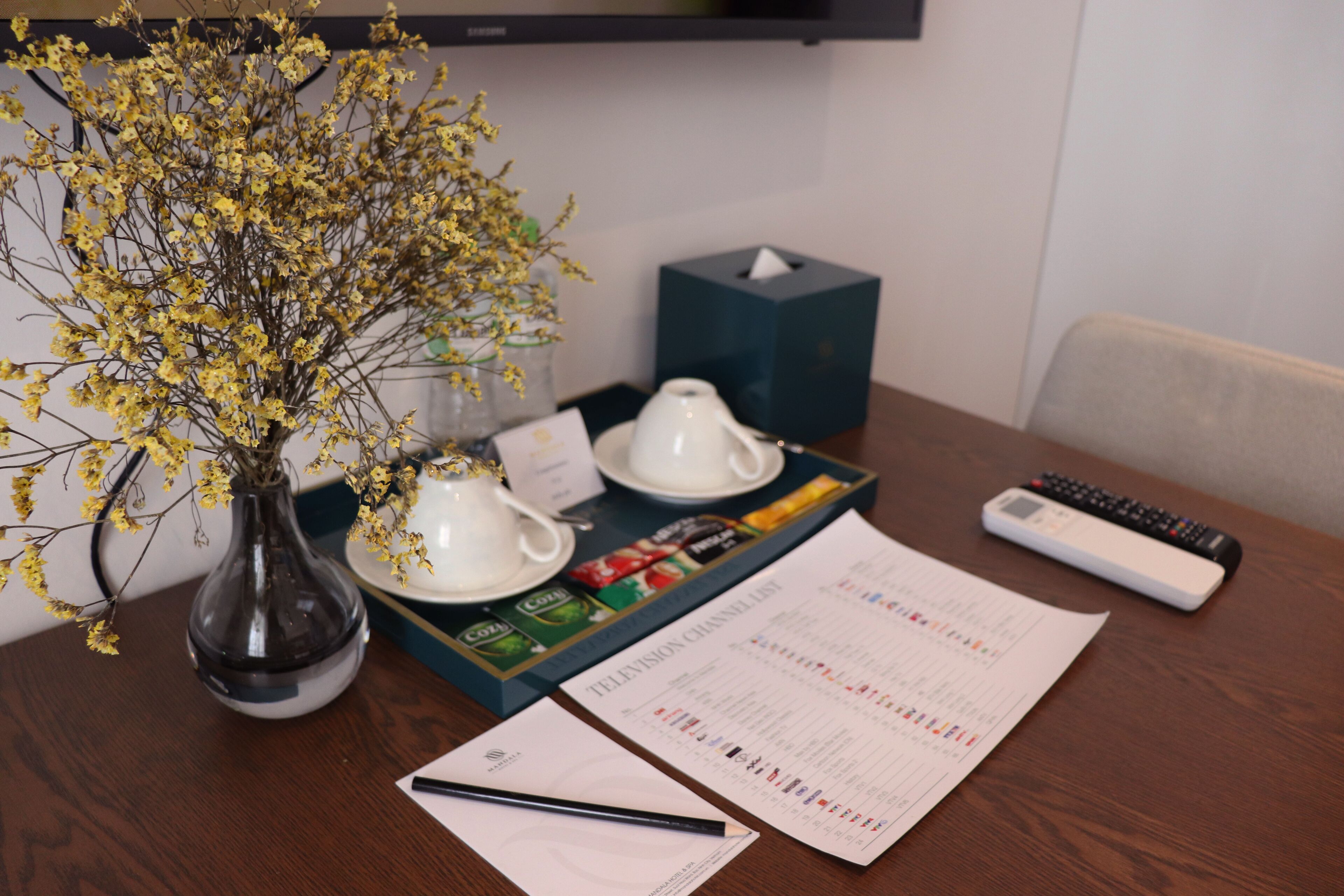 A wooden table with a vase of yellow flowers, two white teacups, a tissue box, a remote control, and a document with a pen.