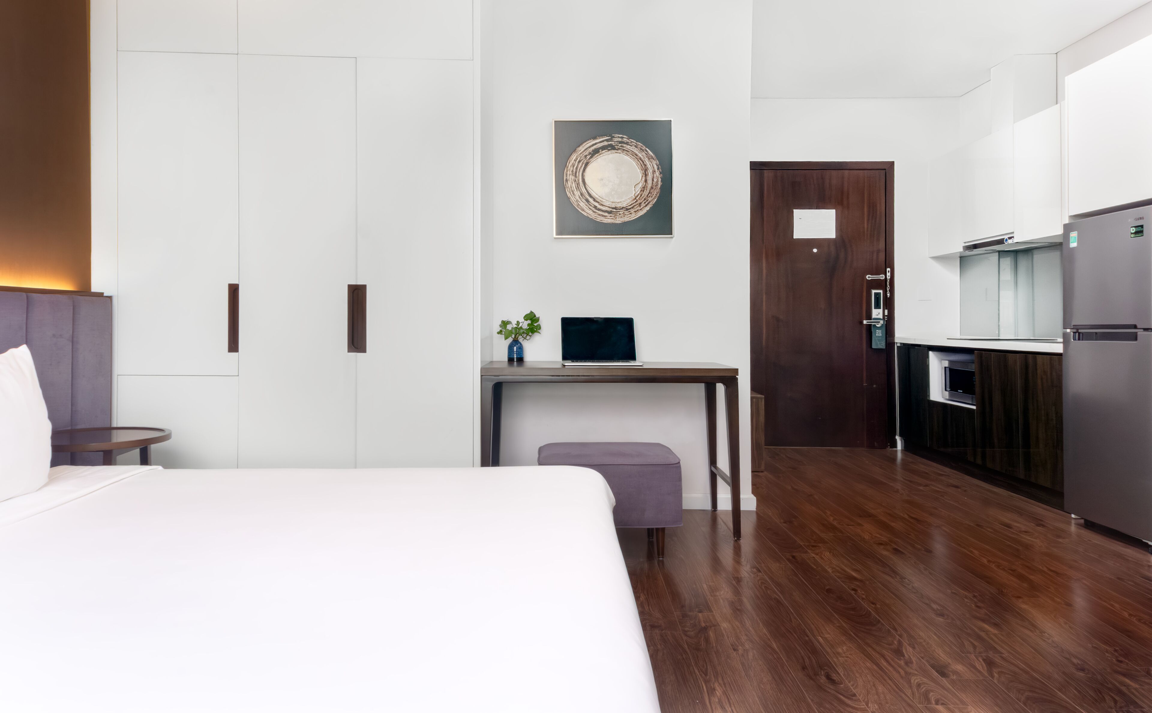 Ett modernt hotellrum med en säng, ett skrivbord med en bärbar dator, en liten växt, en dörr och ett litet pentry.