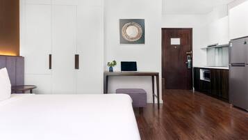 Ett modernt hotellrum med en säng, ett skrivbord med en bärbar dator, en liten växt, en dörr och ett litet pentry.