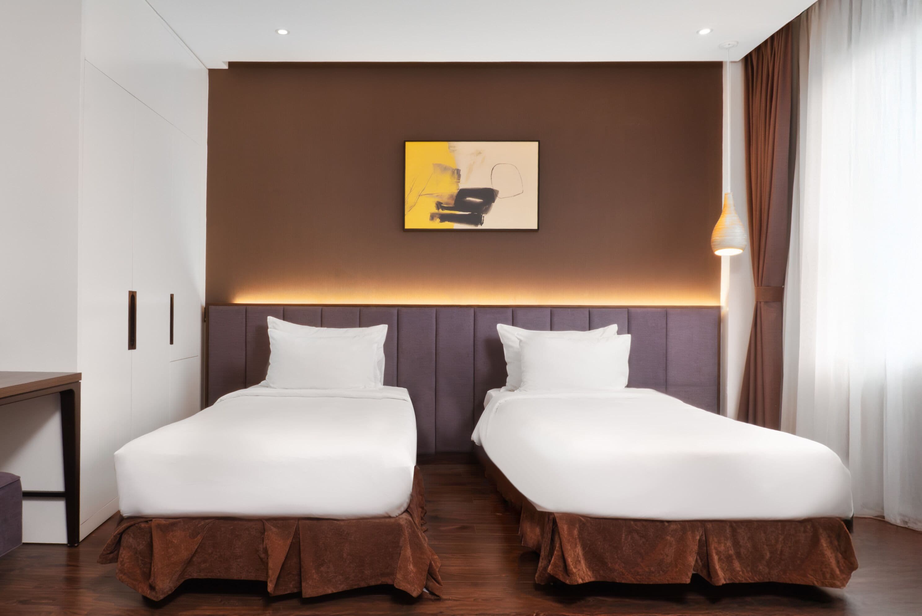 Et hotelværelse med to senge, et skrivebord, en lampe og et maleri på væggen.