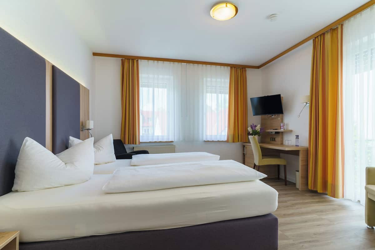 Habitación de hotel con dos camas, un escritorio, televisión y ventanas amplias con cortinas.