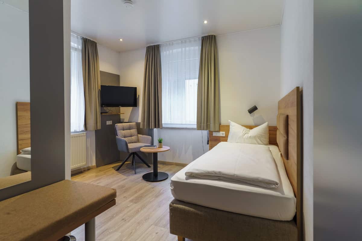 Habitación de hotel con cama, una silla, una mesita, televisor y ventana con cortinas.