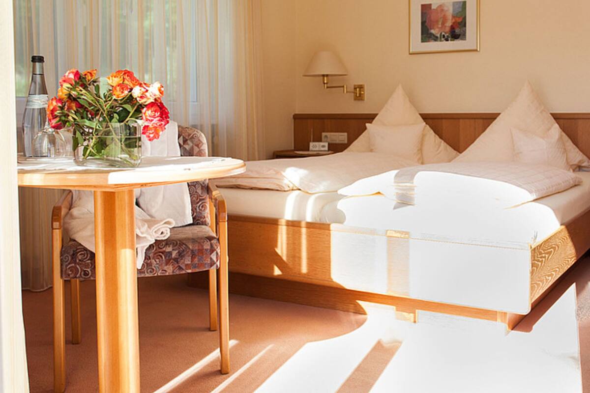 A hotel room with a bed, bedside table, chair, and a vase of flowers.