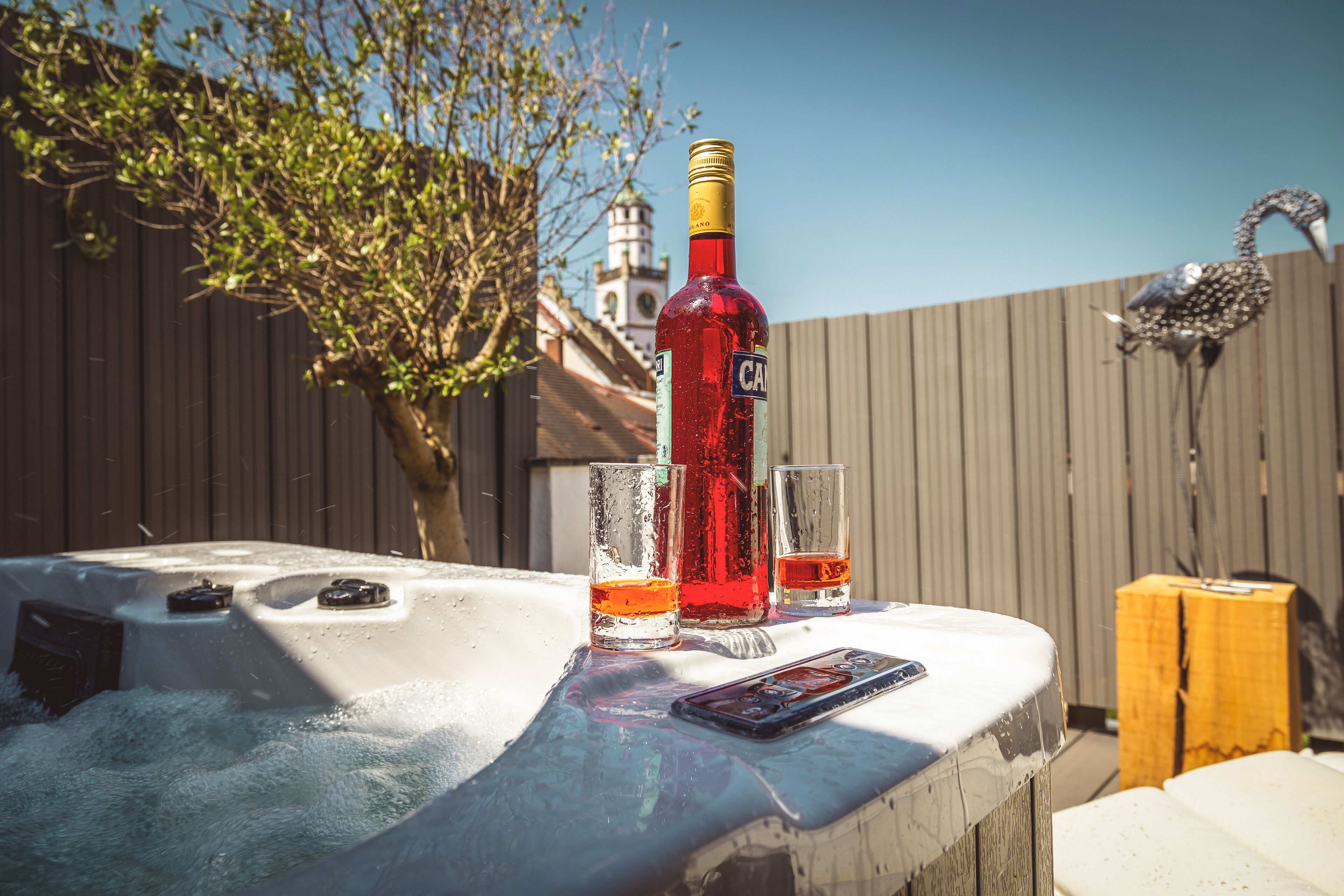 A hot tub with a bottle and two glasses, a wooden fence, and a decorative flamingo statue in the background.