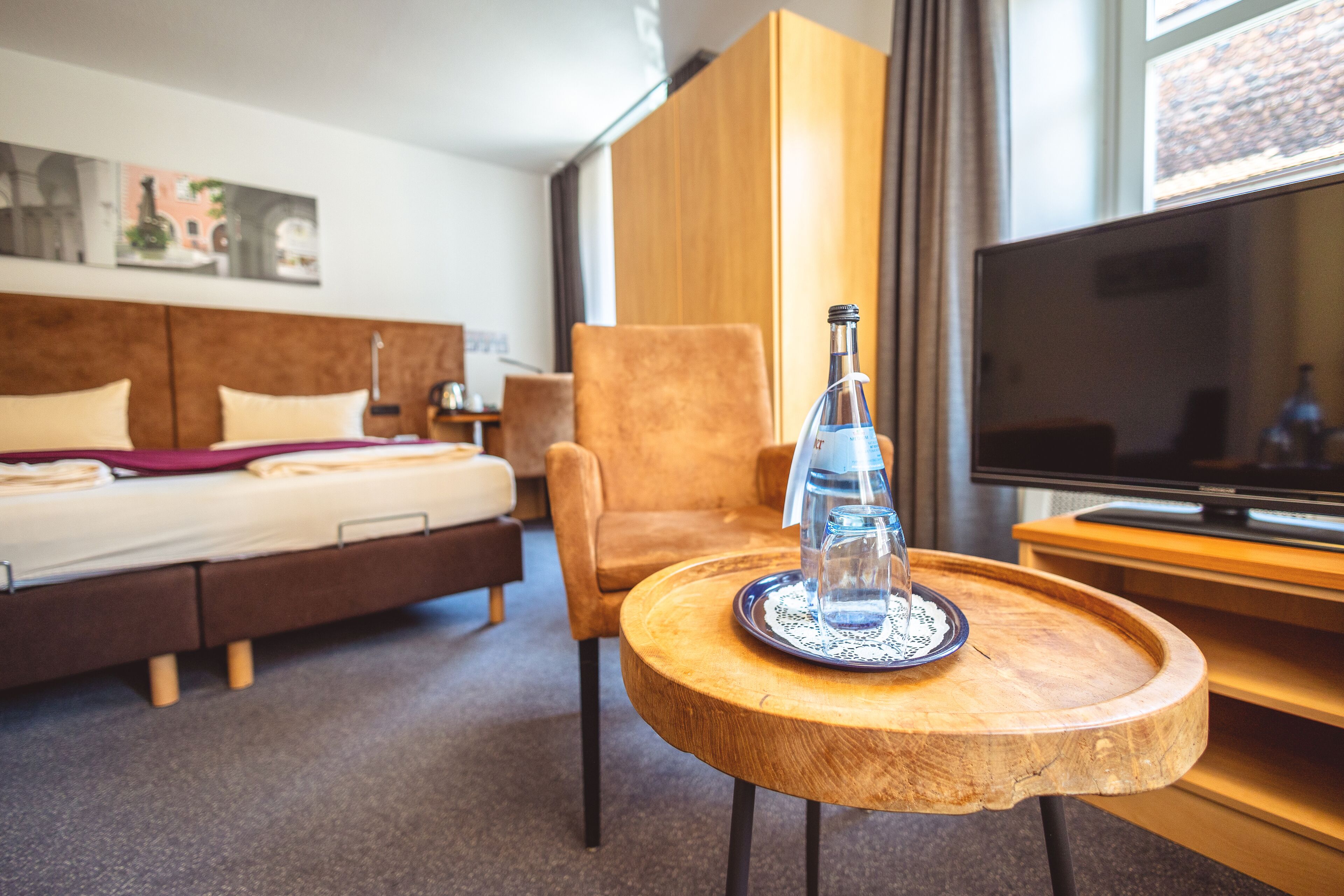A hotel room with a bed, a wooden chair, a small wooden table with a bottle and glasses, and a TV.
