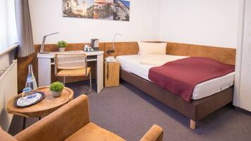 Quarto de hotel com cama, escrivaninha com luminária, cadeira, mesinha com planta e janela com cortinas.
