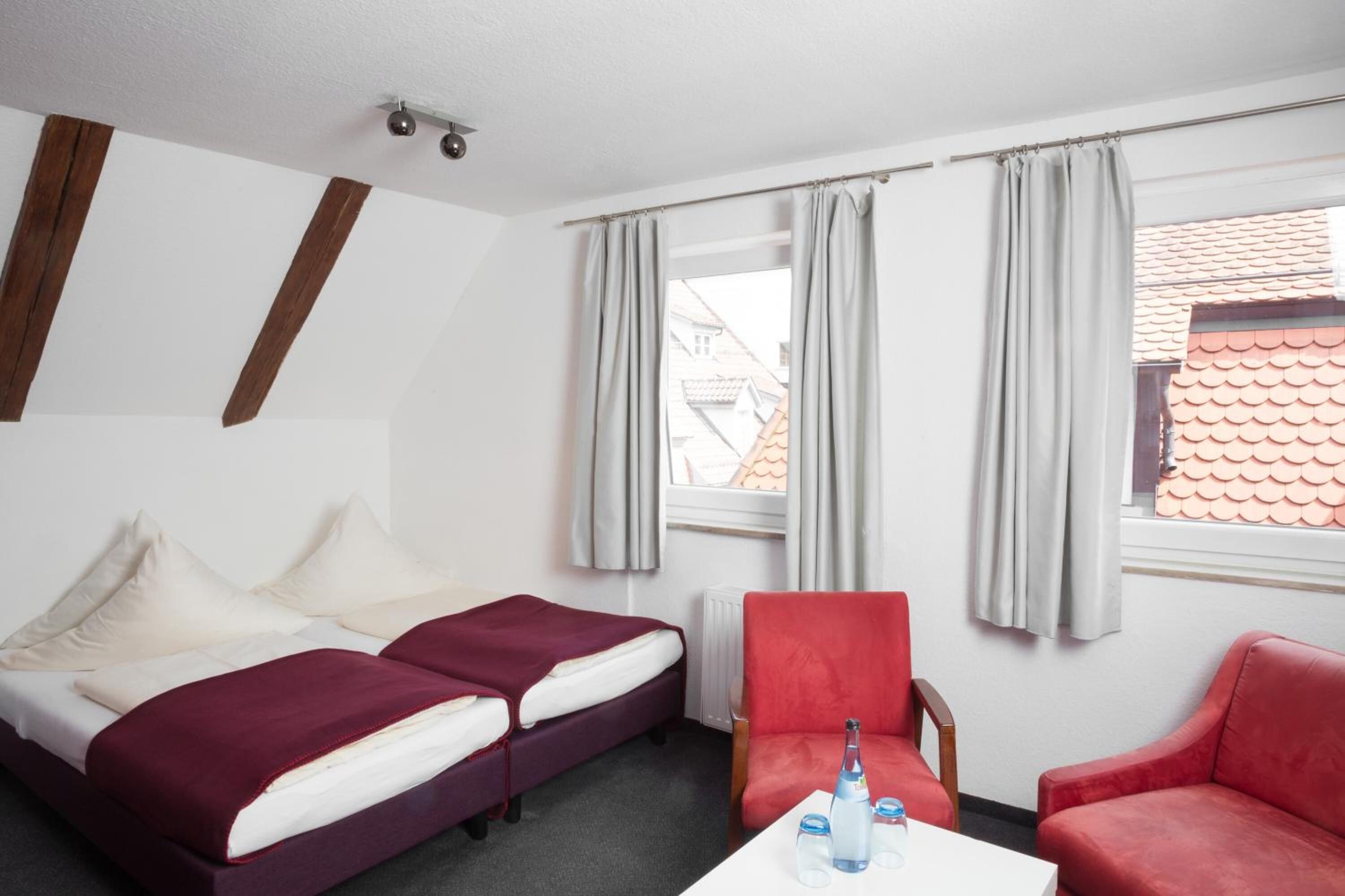 Habitación de hotel con dos camas, un sillón rojo, una mesa blanca con una botella y vasos, y un ventanal con cortinas.