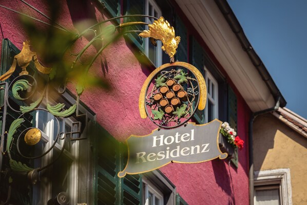 Un letrero de hotel con el nombre 'Hotel Residenz' y elementos decorativos, incluyendo una hoja dorada y un emblema circular con un festón.