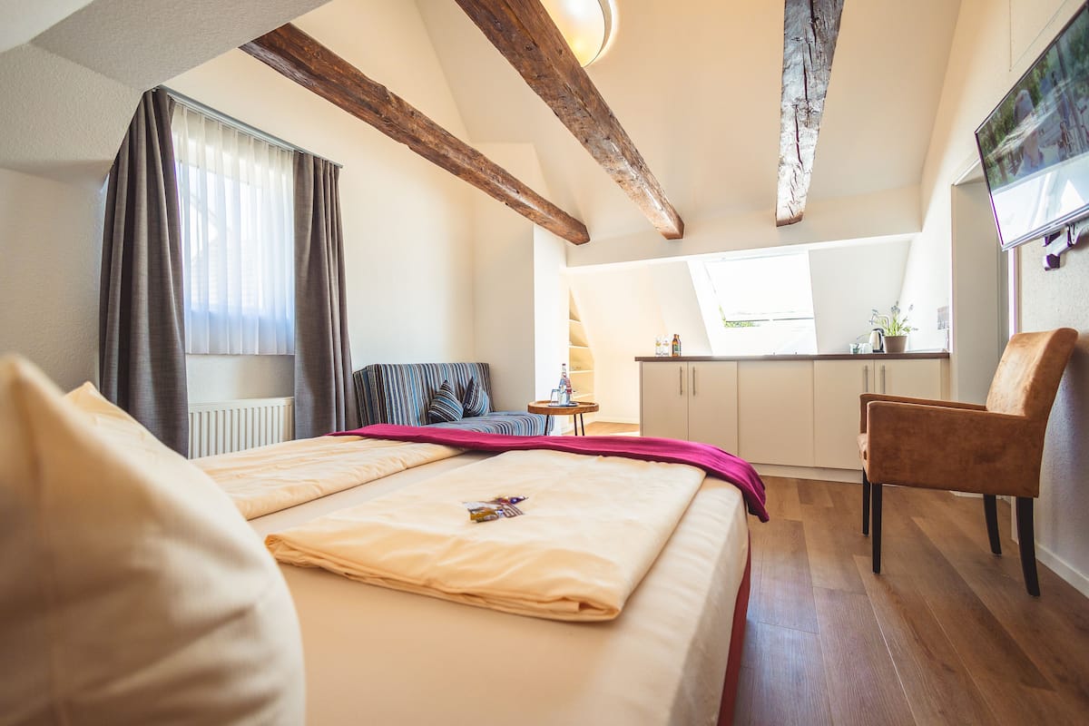 Hotellihuone, jossa on sänky, tuoli, pieni pöytä, ikkuna verhoilla, pieni keittiö ja seinälle asennettu televisio.
