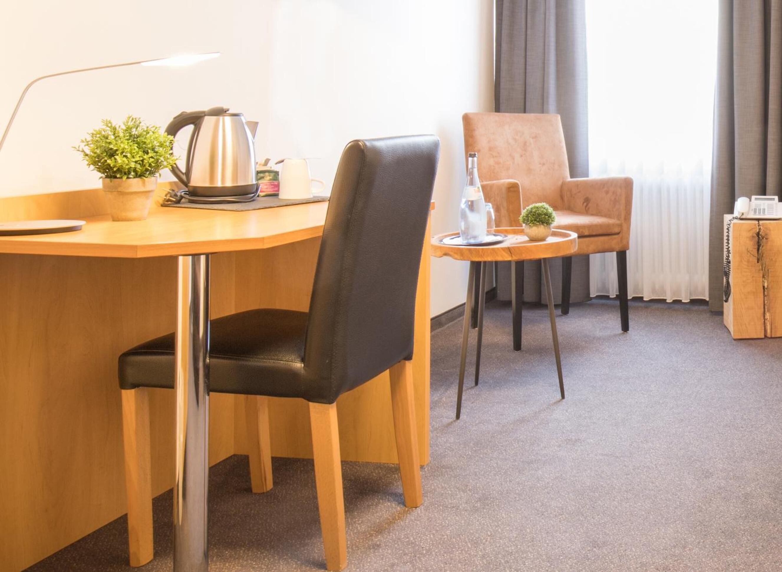 A hotel room with a wooden desk, a black leather chair, a small round table with a plant, a coffee maker, and a telephone.