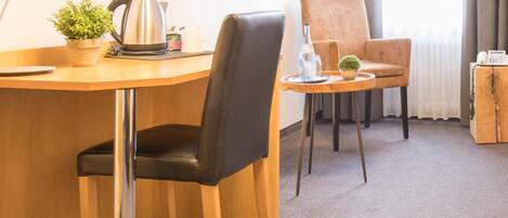 Habitación de hotel con un escritorio de madera, una silla de cuero negro, una mesita redonda con una planta, una cafetera y un teléfono.