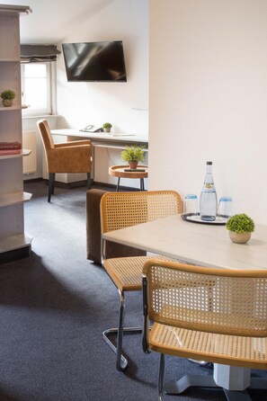 Una habitación de hotel moderna con una mesa redonda grande, dos sillas de mimbre, un televisor montado en la pared y una mesita de noche con una planta en maceta y vasos.
