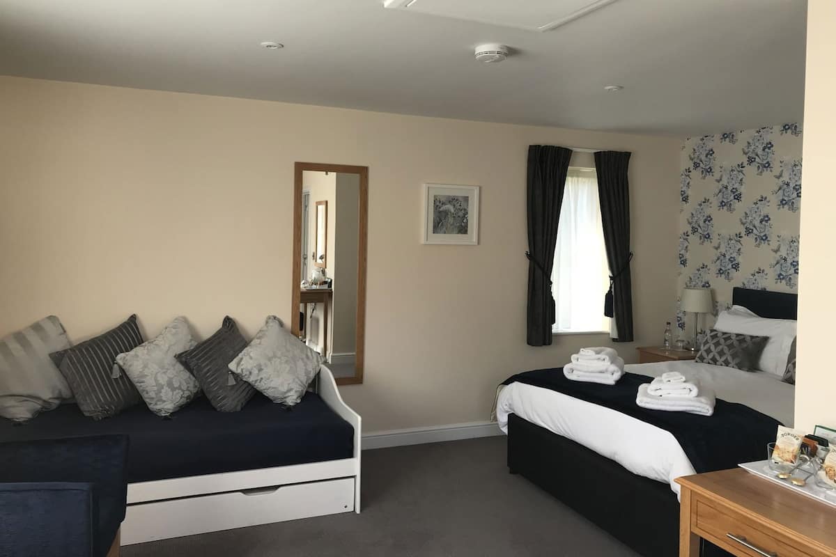Quarto de hotel com cama, sofá, espelho, janela com cortinas, mesa de cabeceira e parede com papel de parede floral.