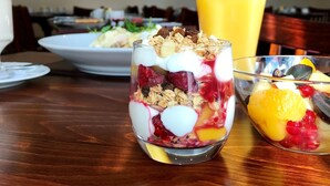 A dining table with a glass of fruit salad, a bowl of cereal, and a glass of orange juice.
