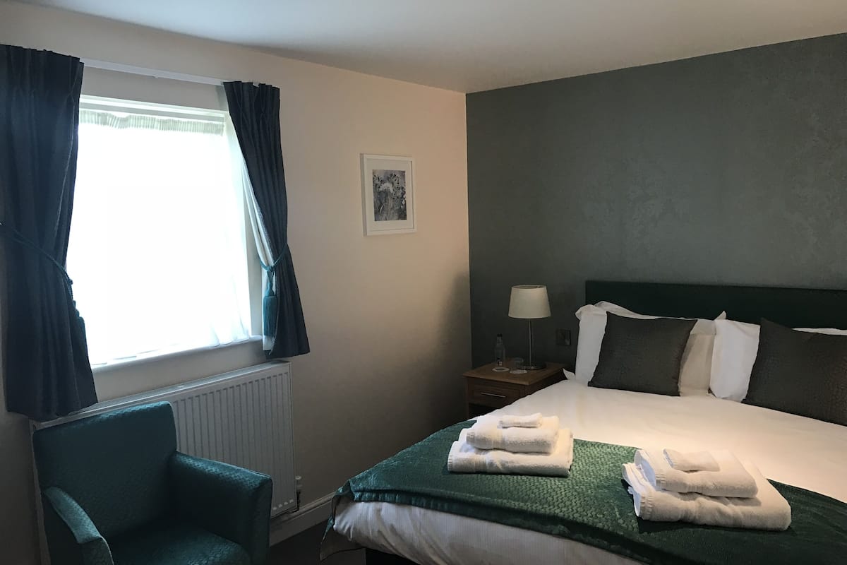 Quarto de hotel com uma cama, uma cadeira, uma mesa de cabeceira com abajur, um quadro na parede e uma janela com cortinas.
