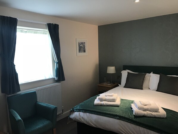 A hotel room with a bed, a chair, a nightstand with a lamp, a framed picture on the wall, and a window with curtains.