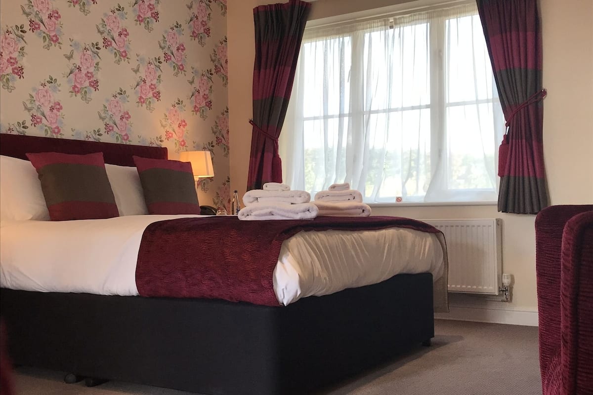 Quarto de hotel com uma cama grande, uma mesa de cabeceira com abajur e uma janela com cortinas.