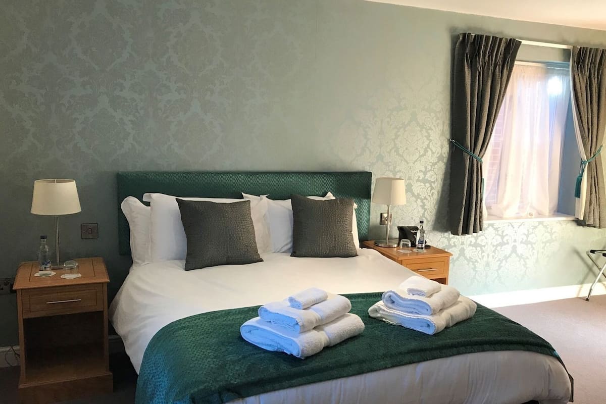 Cama bem arrumada com almofadas, manta verde e toalhas dobradas no painel da cabeceira. Há duas mesas de cabeceira com abajures e uma janela com cortinas.