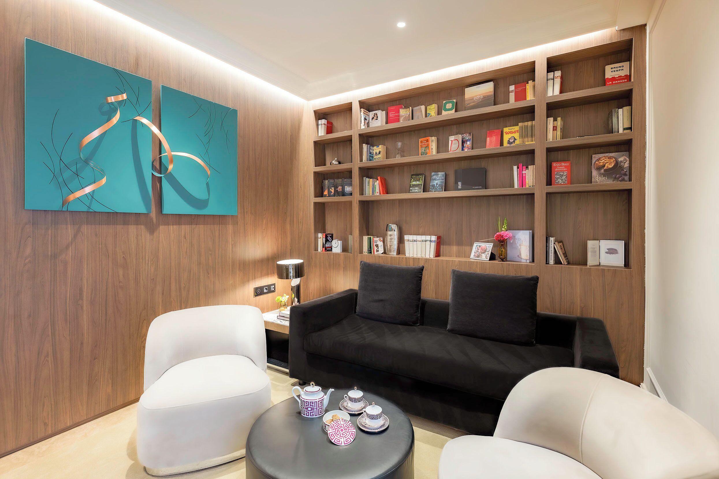 Una sala de estar moderna con un sofá negro, sillones blancos, una mesa de centro con teteras y una estantería llena de libros y objetos decorativos.