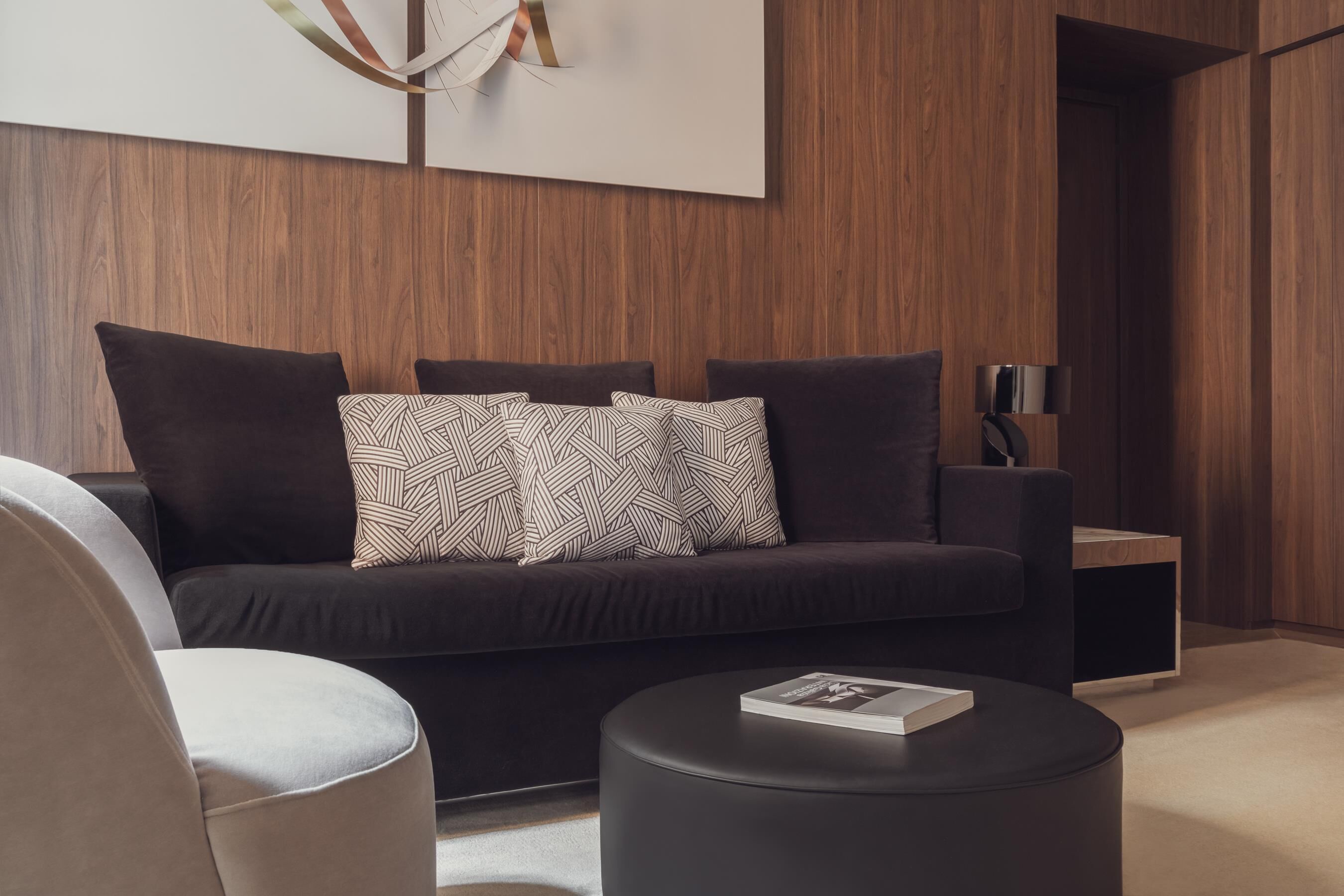 Un salon moderne avec un canapé de couleur foncée, des coussins à motifs, un pouf rond noir et une table d’appoint avec une lampe.